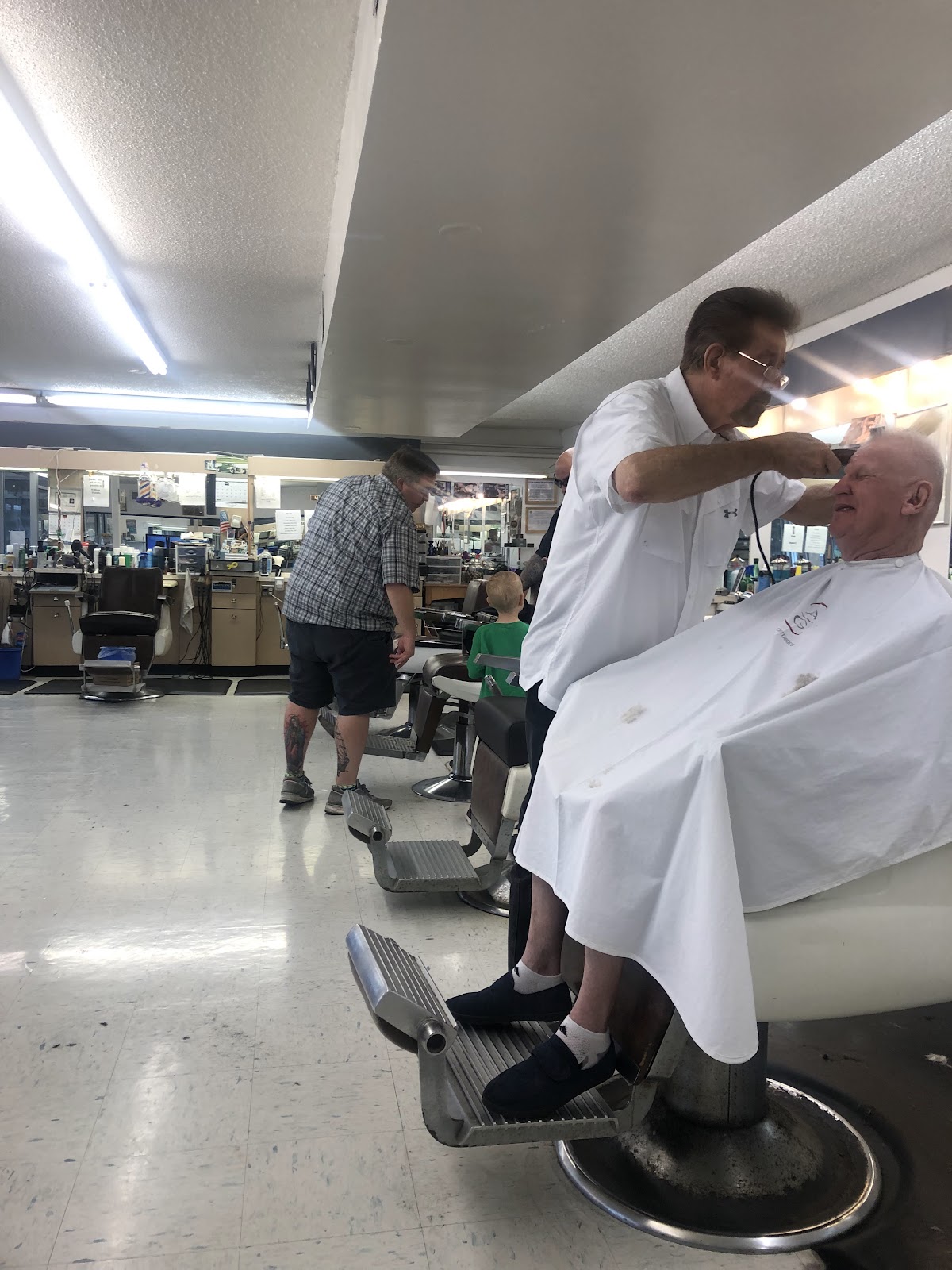 A barber cutting a seated customer's hair with a child visible in the background